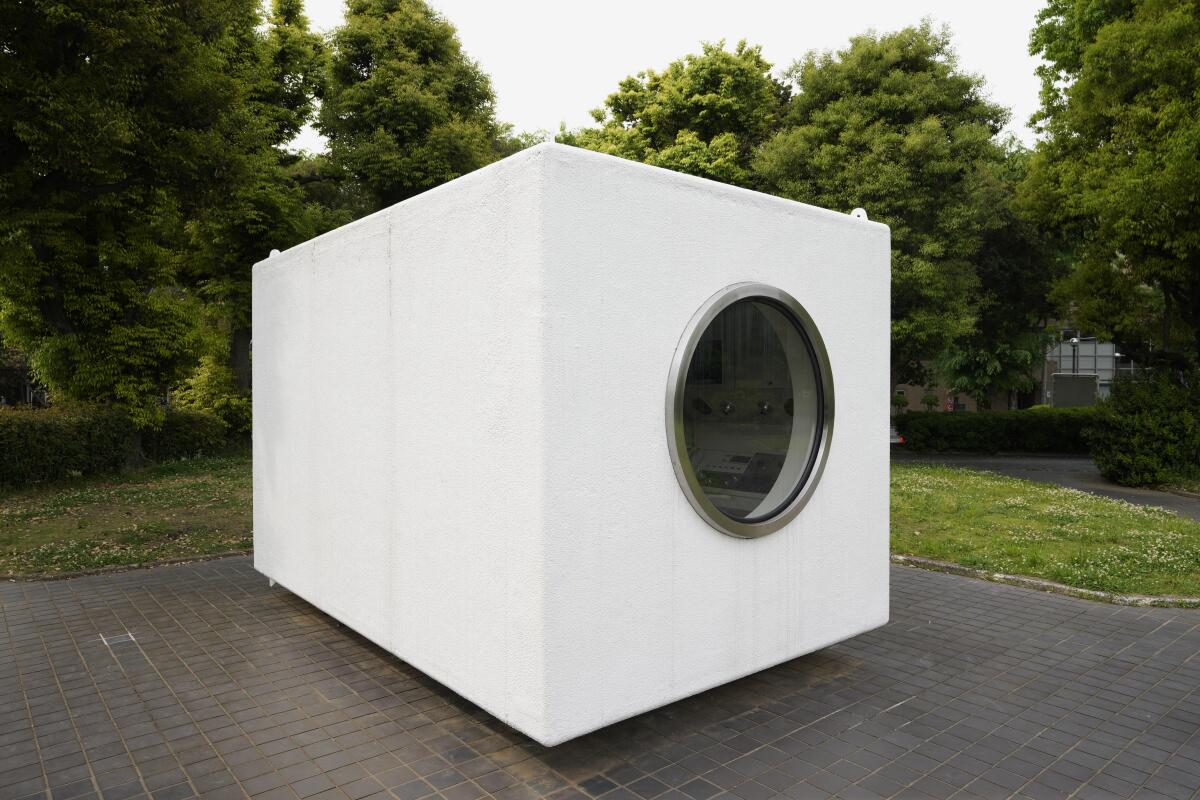A white capsule apartment with a circular window is displayed in a garden space.