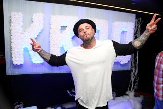 Musician Shifty Shellshock wears a black hat and white t-shirt and makes peace signs with his outstretched hands