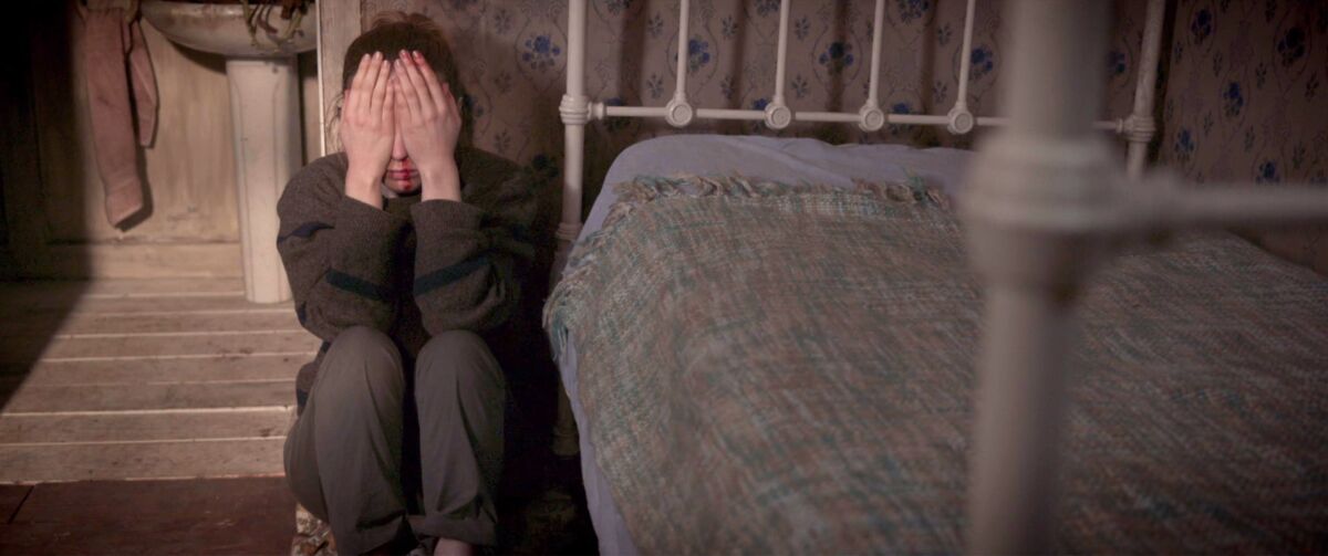 A woman covers her eyes with her hands while sitting on the floor next to a bed.