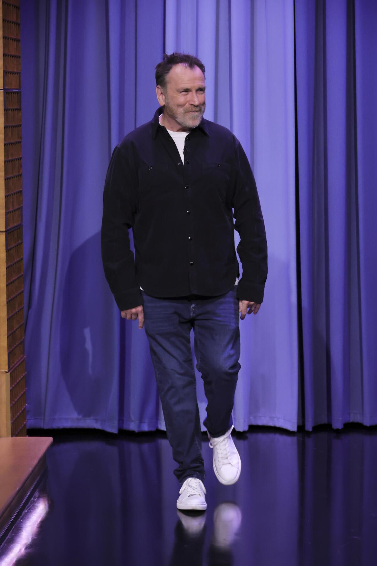 A man in jeans and a dark jacket walks onto a stage.