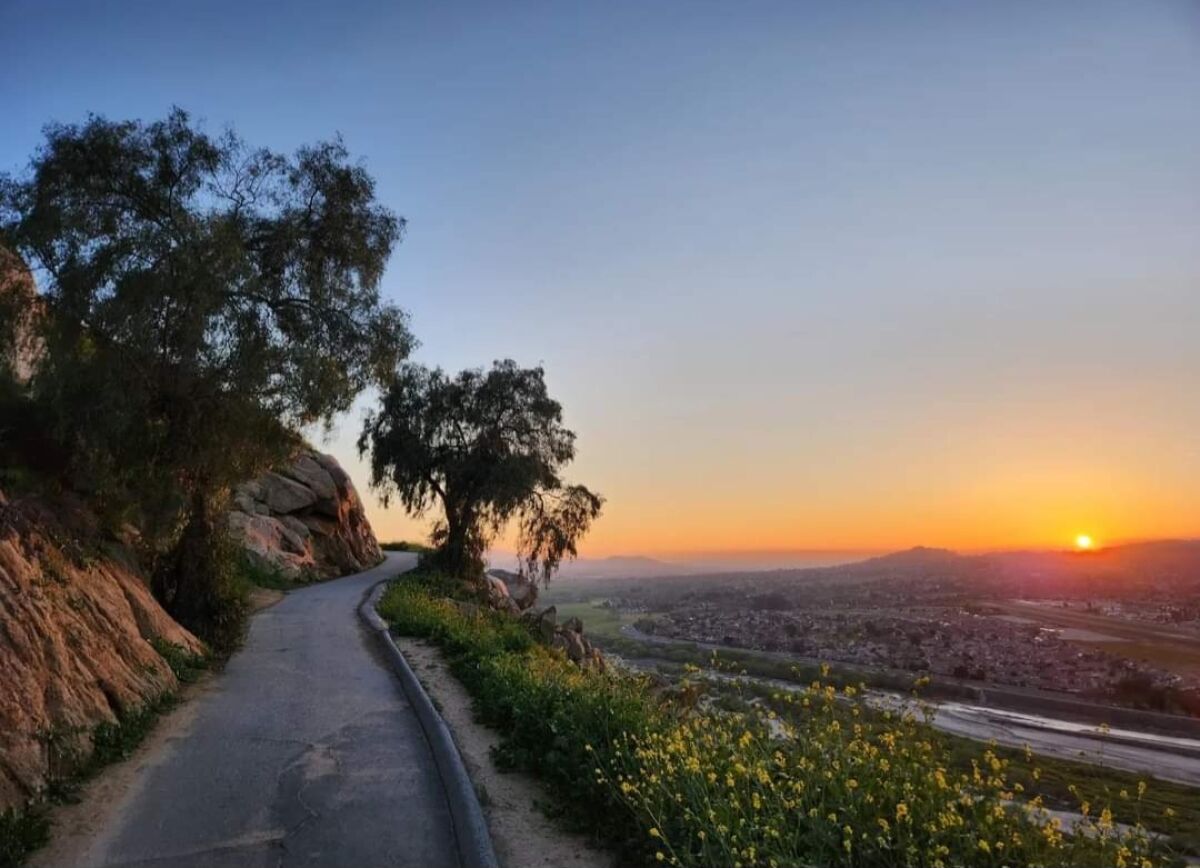 A hiking trail winds along a mountain side with a sunset and wide view of a city shown to the right.