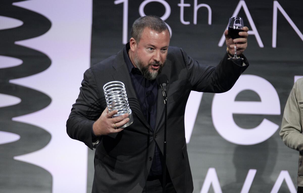 A man in a black suit accepts an award and raises a glass of wine