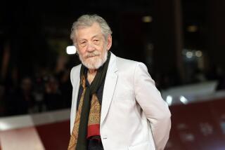 Ian McKellen with facial hair in a white blazer and a black shirt and colorful scar posing with his hands behind his back