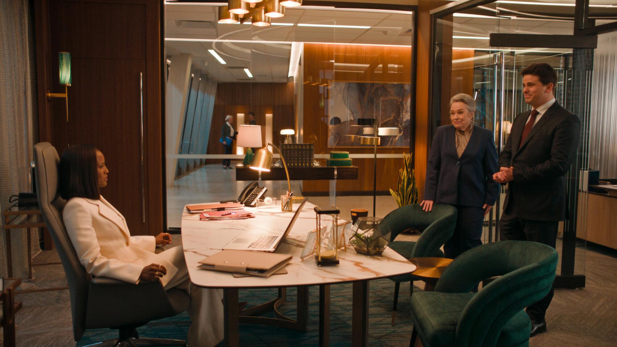 A woman in a white suit sits behind a desk and looks at a woman and a man.