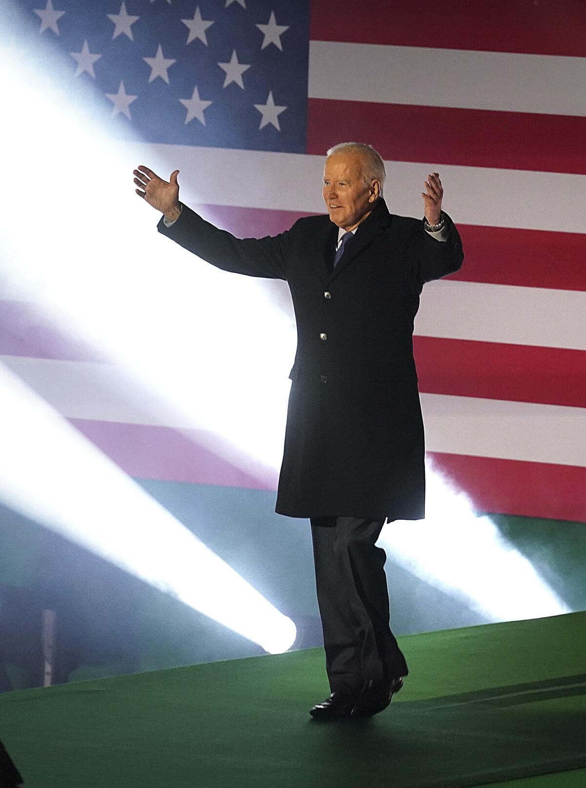 Joe Biden, with his arms outstretched, walks onto a stage with a large American flag as a backdrop