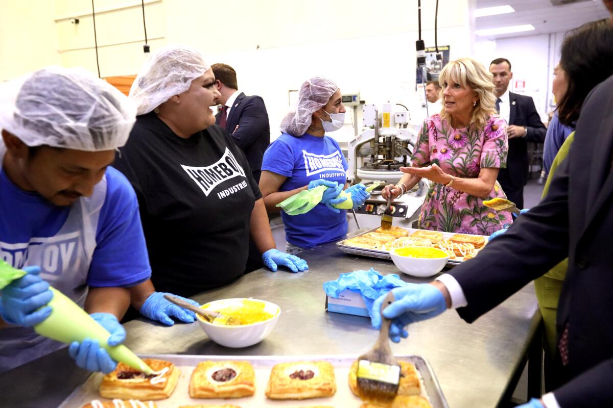 First Lady Jill Biden speaks with people who are putting glaze on pastries at a bakery table.