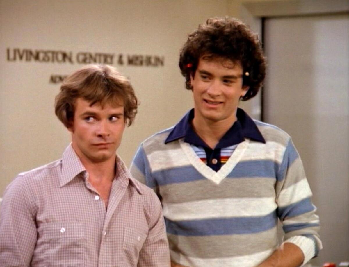 Peter Scolari in a pink, checkered shirt stands next to Tom Hanks in a striped shirt