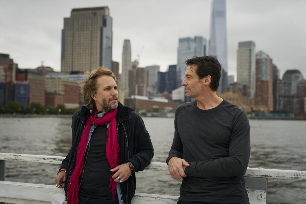 Two men lean against a railing with water and tall buildings in the background.