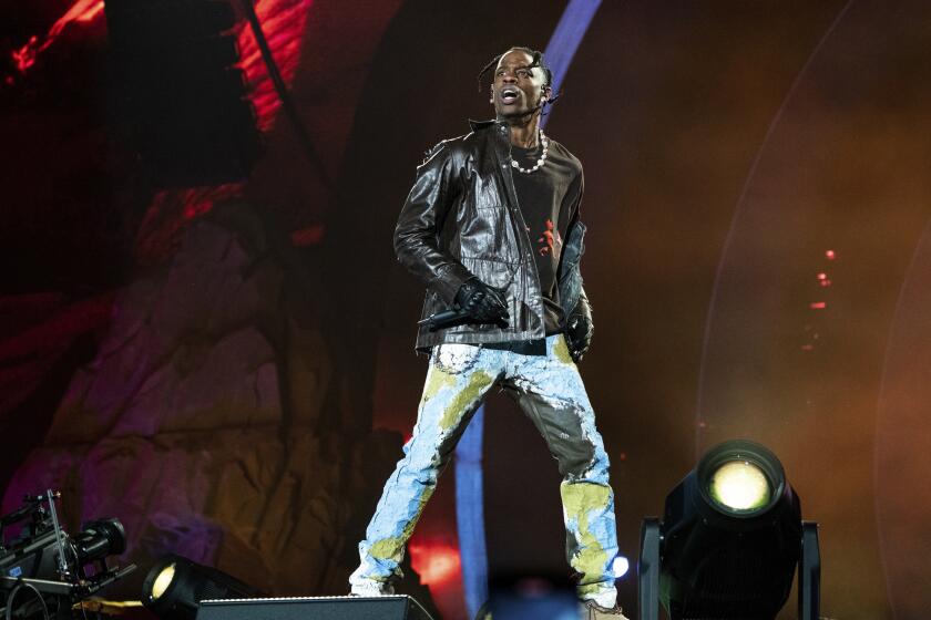 Travis Scott in a black leather jacket, jeans with a chain standing on a stage and looking over his right shoulder