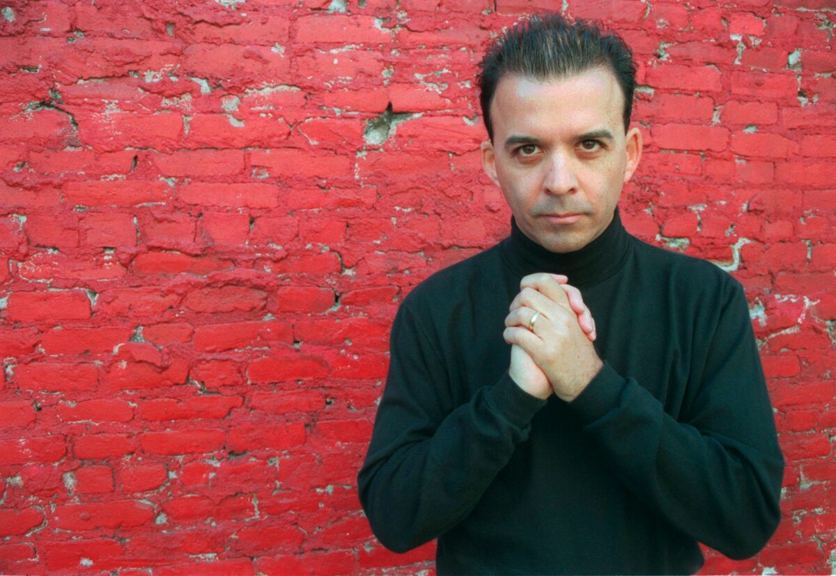 A man in a black turtleneck clasps his hands before a red brick wall.