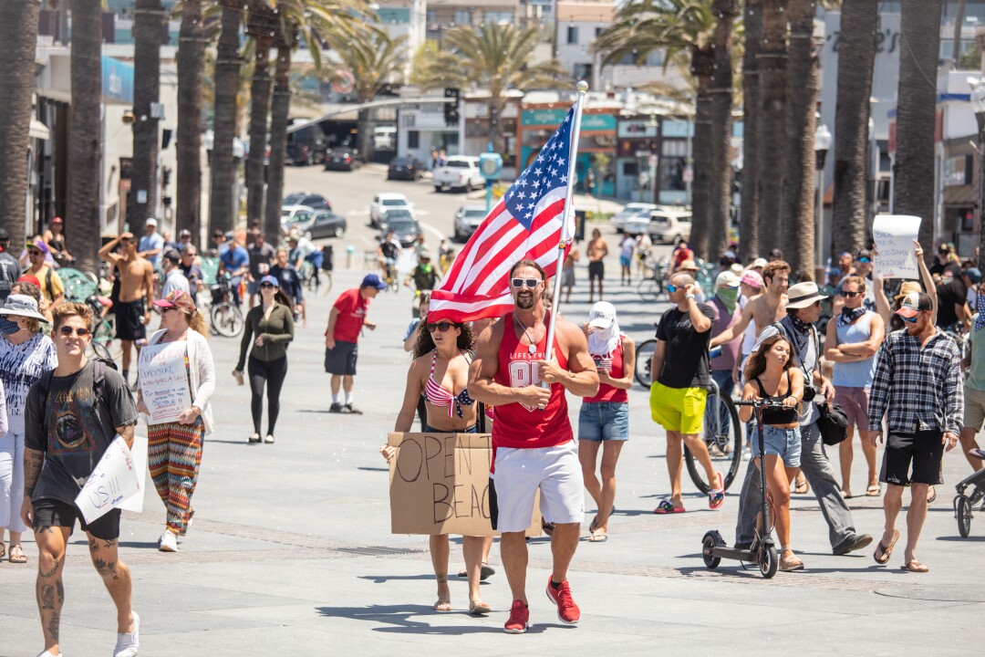 Hermosa Beach residents Aaron Reed, with an American flag and Ashley Puida, center-left, brought their protest to Pier Plaza in Hermosa Beach and were joined by about 100 people in chanting to “open the beach.” 