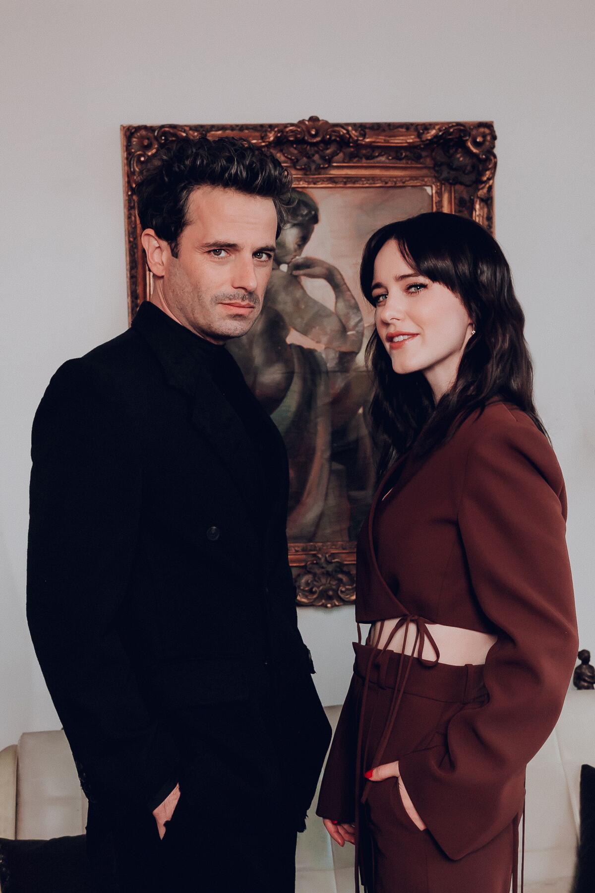 A man and a woman stand in front a painting and look at the camera.