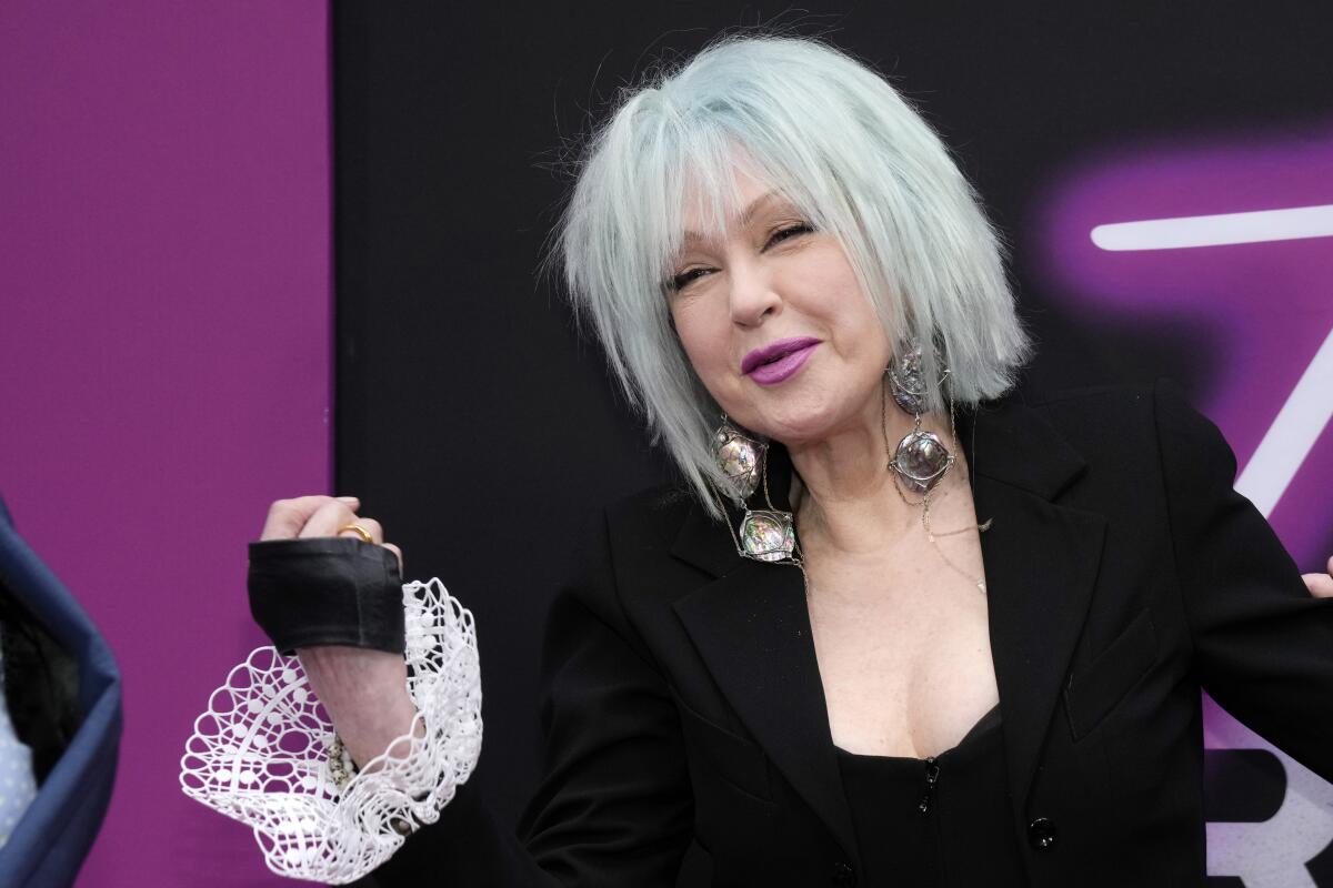 Cyndi Lauper in a pale blue bob, large earrings and a black suit with white ruffle trim on the sleeve