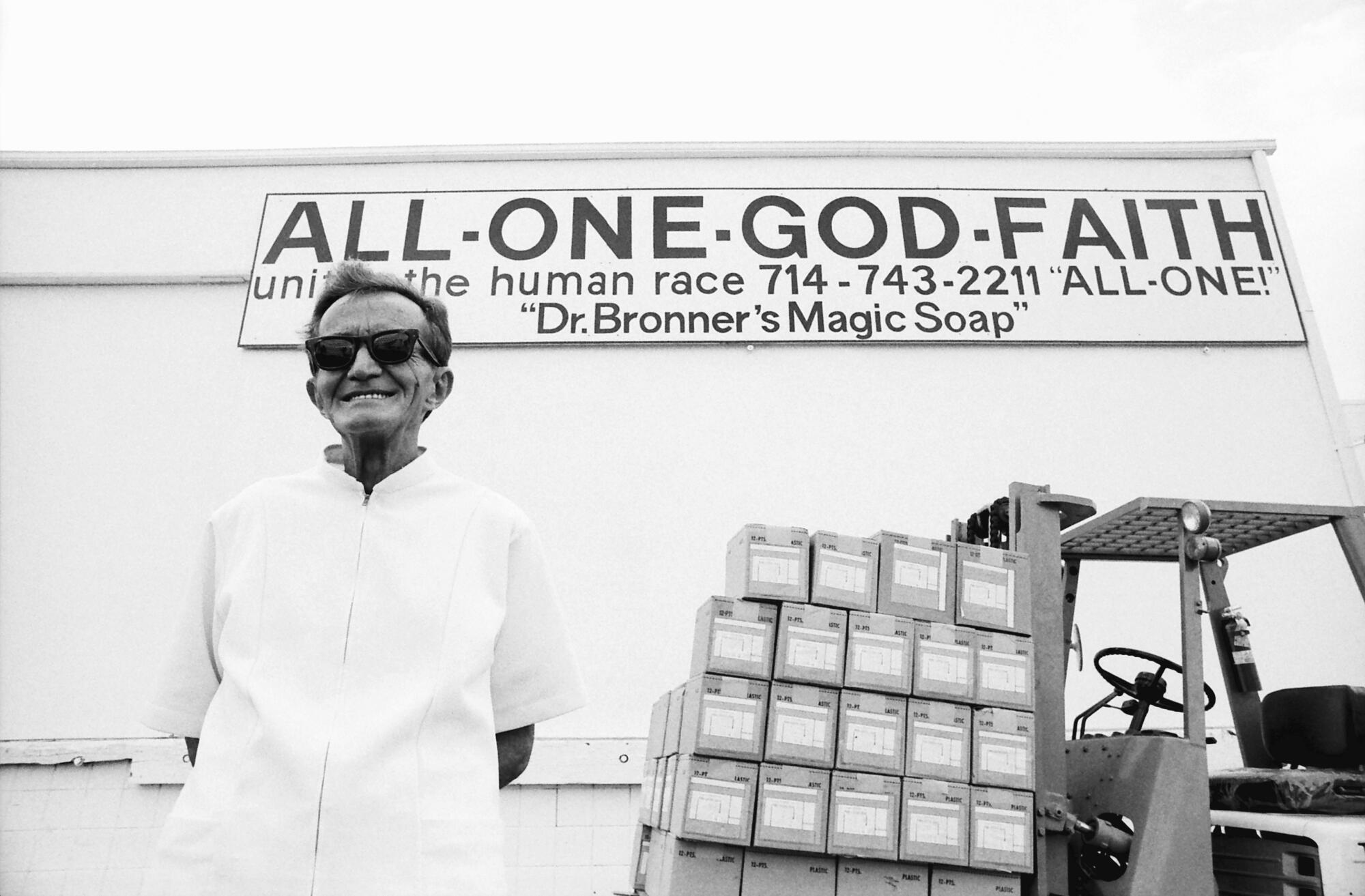 A black-and-white photo of a man by a forklift loaded with boxes, outside a building with a sign reading "All-One-God-Faith"