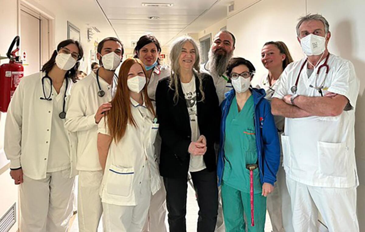 A woman in a black outfit surrounded by eight people in scrubs and white medical gear standing in a hospital hallway