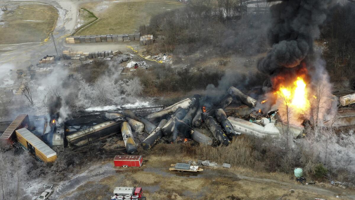Smoke rises from a pile of derailed and charred train cars, with a fire burning at one end 