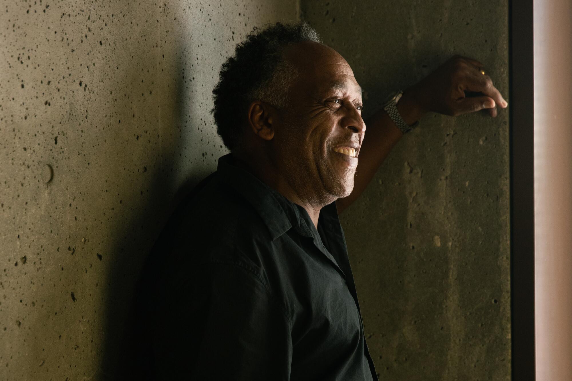 Henry Taylor wearing a black shirt and leaning on a concrete wall, his face illuminated by sunlight