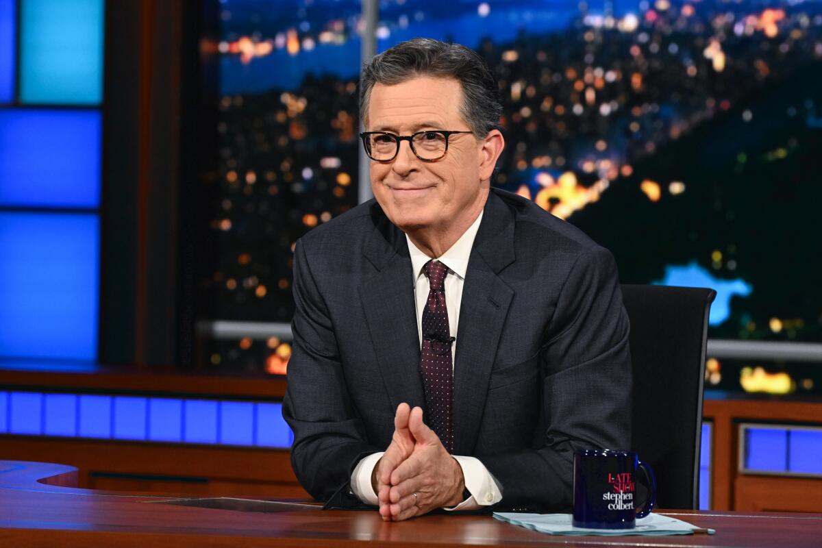 Stephen Colbert folds his hands together and smiles while seated and leaning forward over a desk