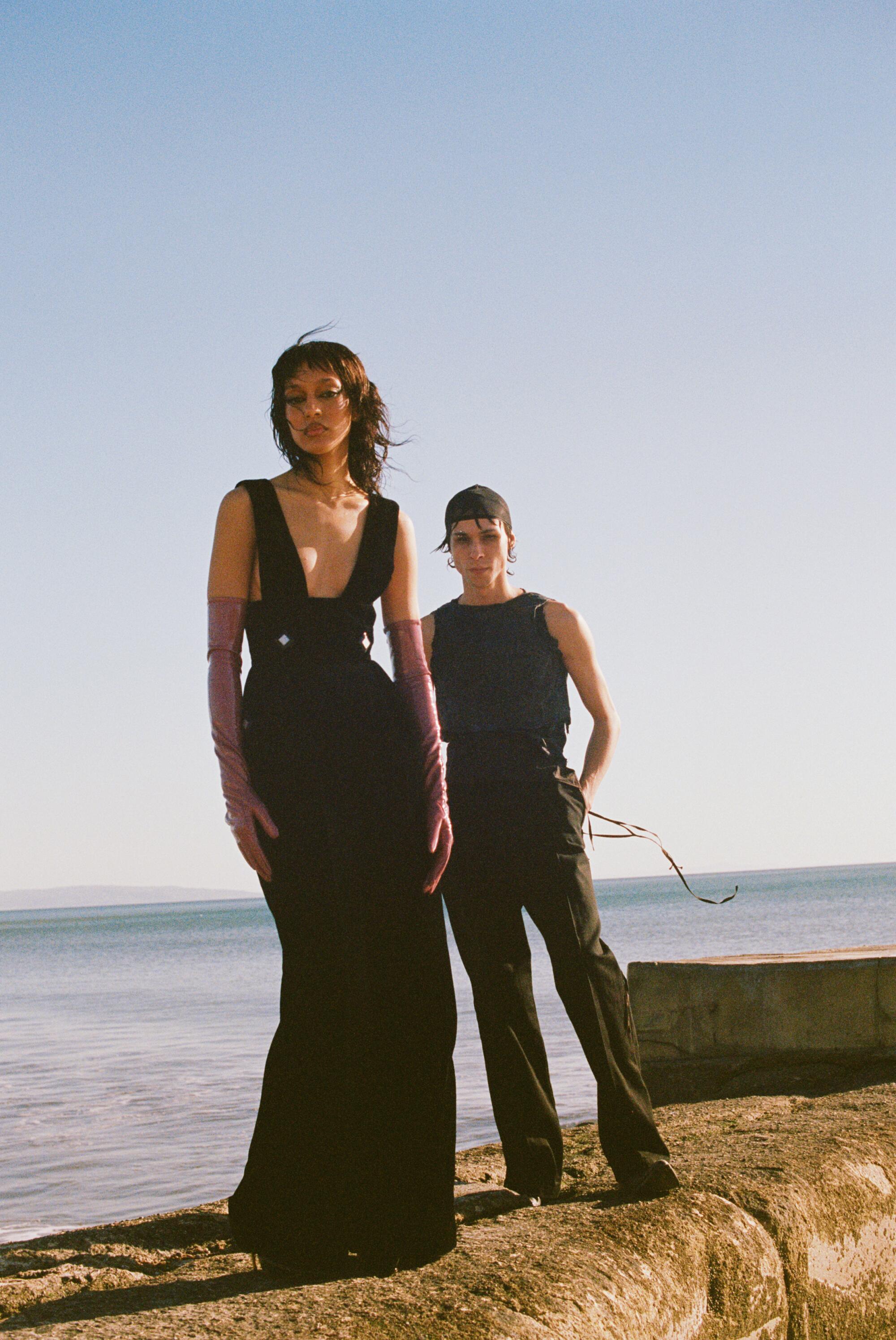 A model in a floor-length black dress and a model in black tank top and slacks stand on a rock wall.