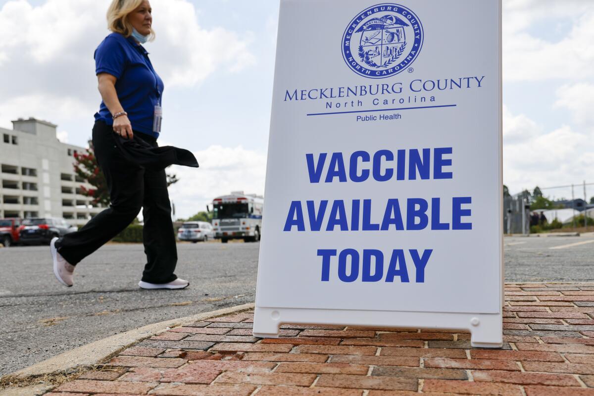 A woman walks by a sign on a sidewalk that says "Vaccine available today."