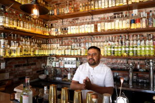 A man stands behind a bar, the wall behind him lined with mezcal bottles.