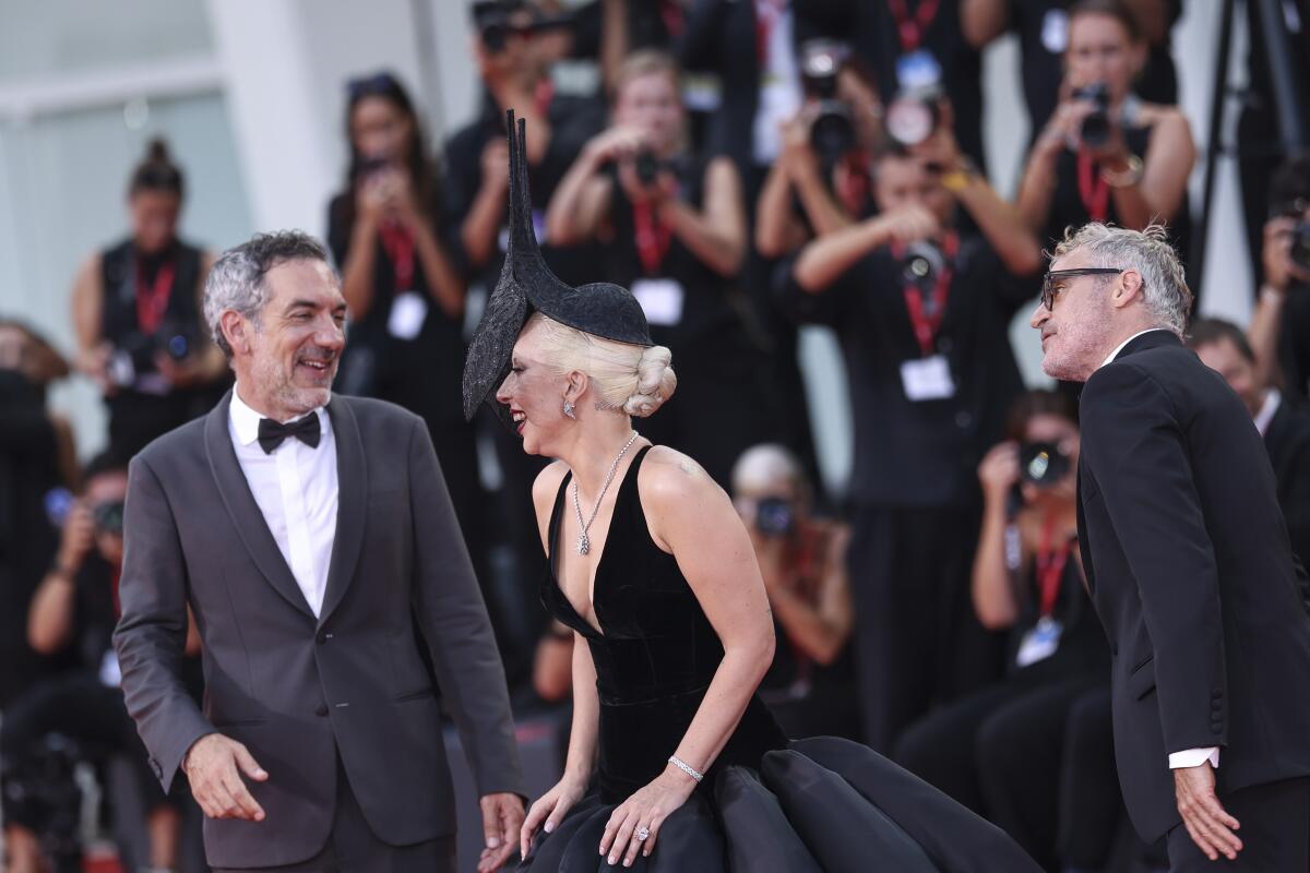 Todd Phillips and Joaquin Phoenix, in tuxedos, flank Lady Gaga, in a black dress and hat, in front of photographers.