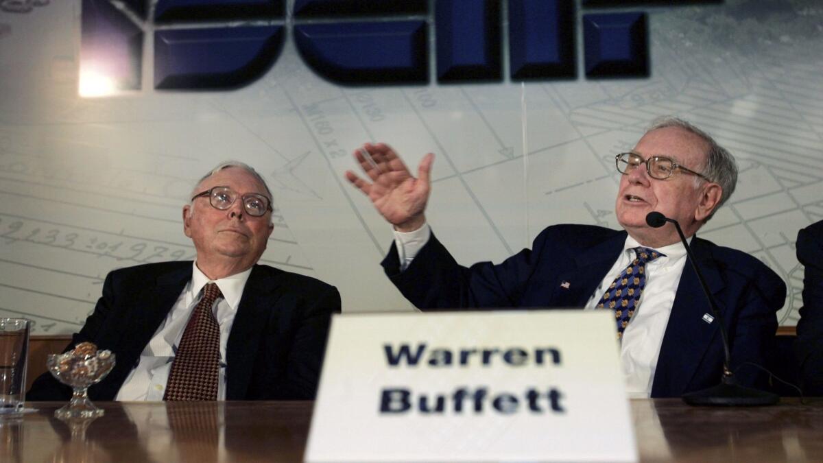 A man behind a "Warren Buffett" name card on a table raises his right hand as a man next to him watches.