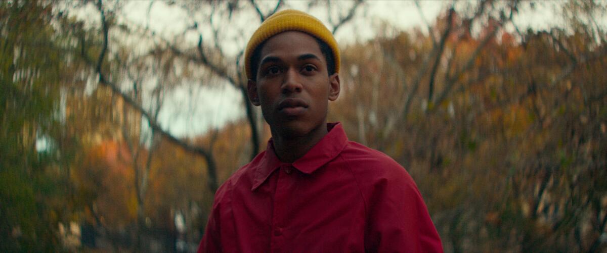 Kelvin Harrison Jr., in a yellow beanie and red shirt, stands in front of trees and fall foliage.