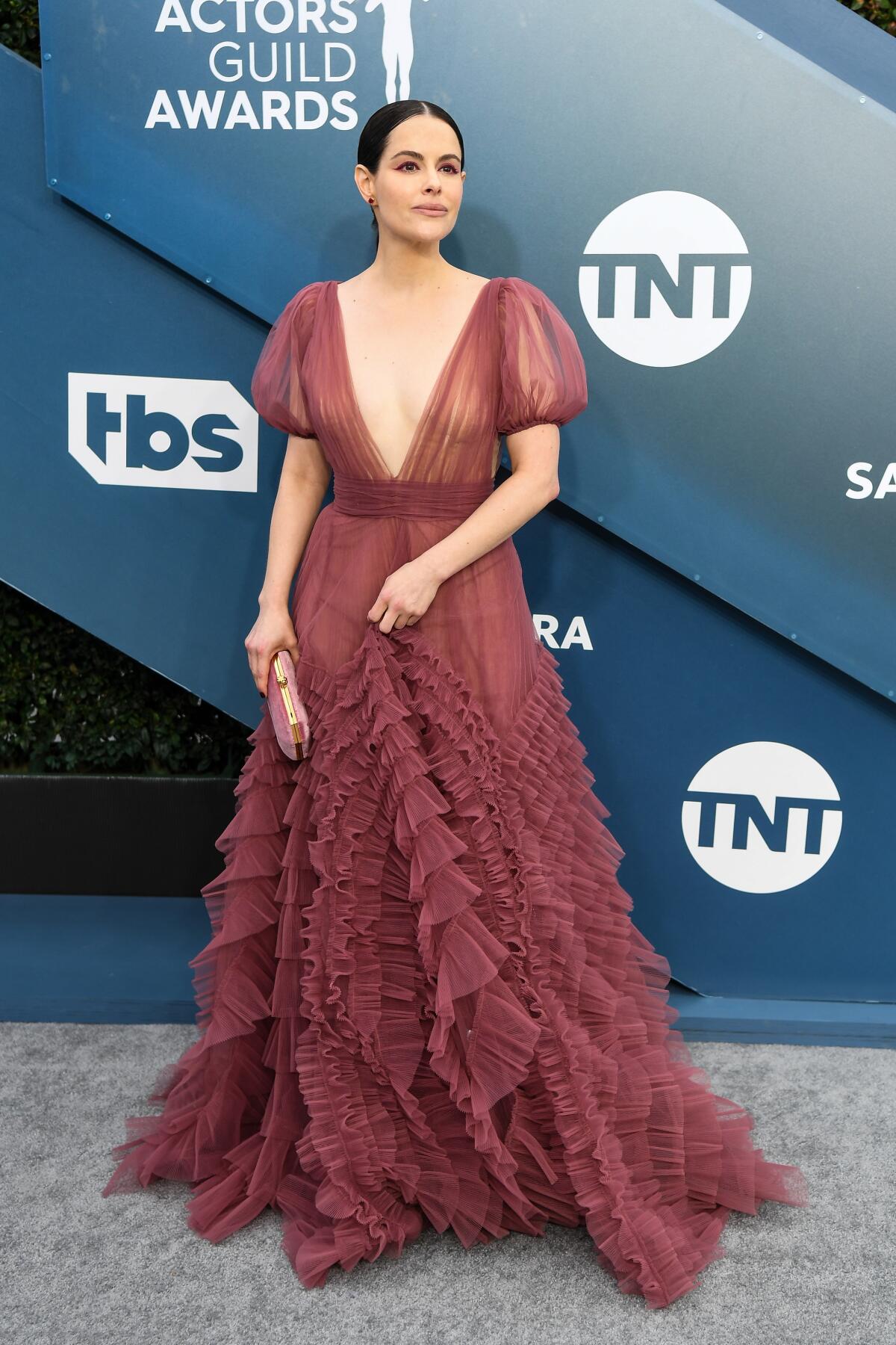 Emily Hampshire arrives at the SAG Awards in a long ruffled gown with a deep V neck in front of a backdrop