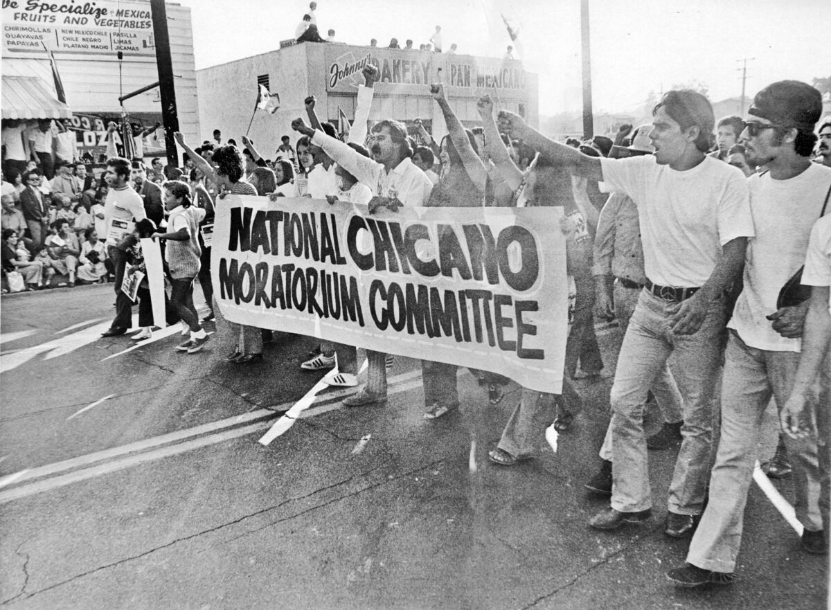 A black and white image from the 1970s shows protestors carrying a sign that reads "National Chicano Moratorium Committee"