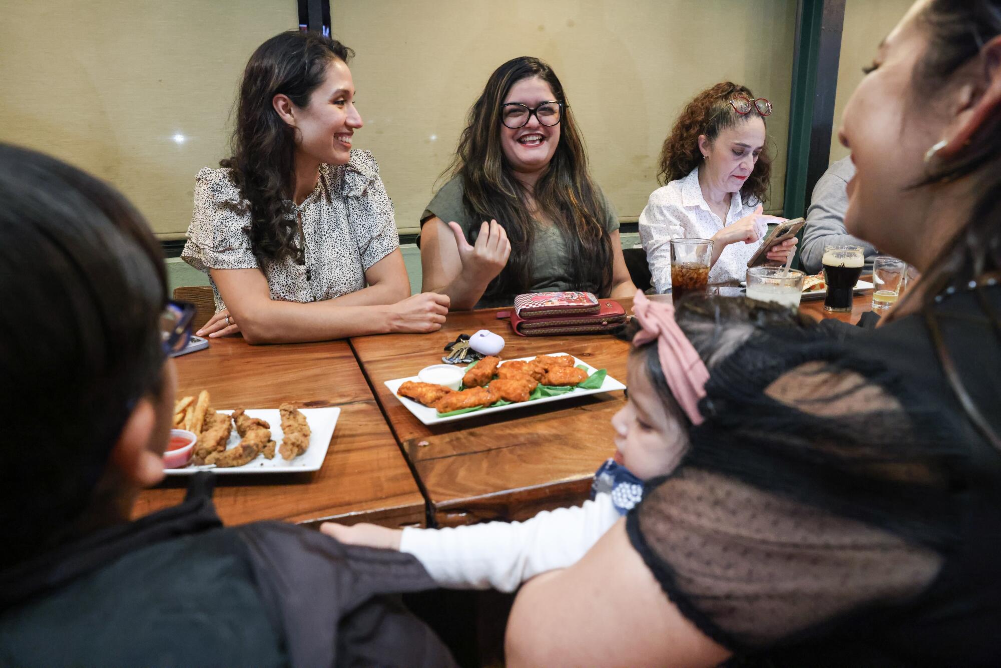 A group of smiling women converse while seated at tables with plates of food in front of them