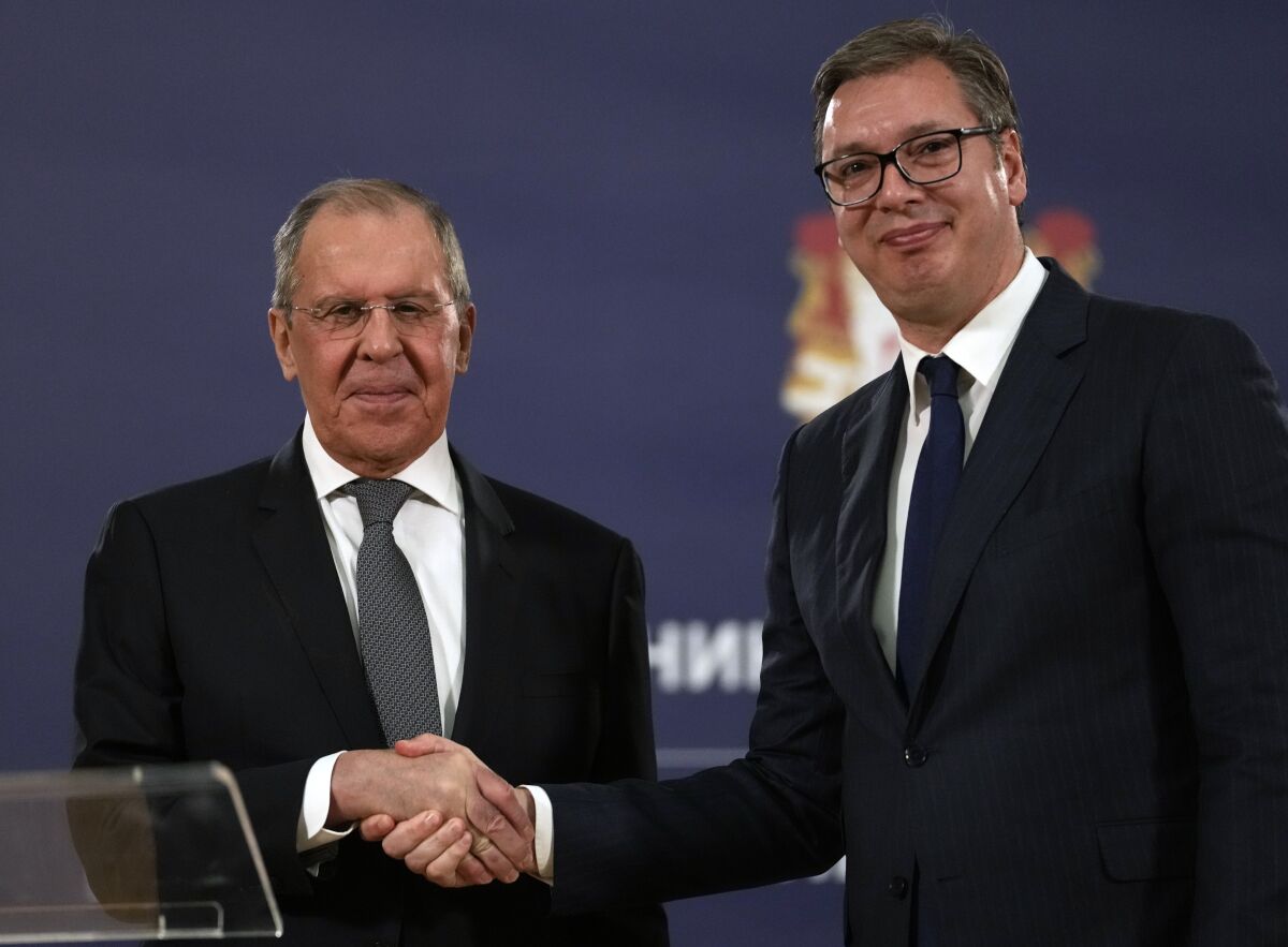 Two men in dark suits smile and shake hands.