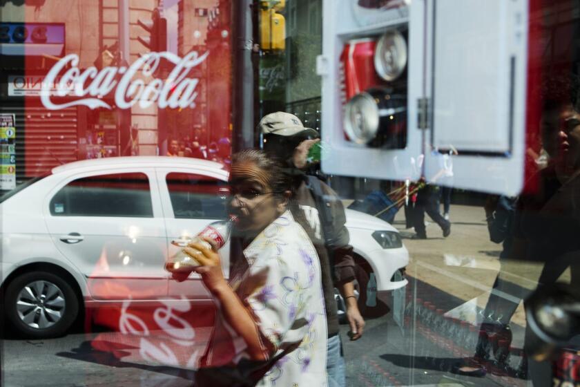 FILE - In this Oct. 9, 2014 file photo, a woman is reflected in a Coca-Cola store window display as she drinks a Coke in Mexico City. Mexicans are among the biggest soda drinkers in the world, so residents of the southern city of Ciudad Altamirano were hit hard when first Coca-Cola then Pepsi closed their distribution centers amid drug gang extortion demands. (AP Photo/Rebecca Blackwell, File)