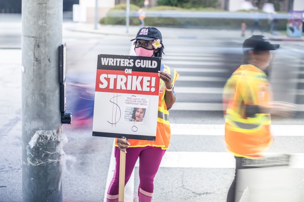A woman wears a pink mask and a baseball cap and carries a writers' strike sign. 