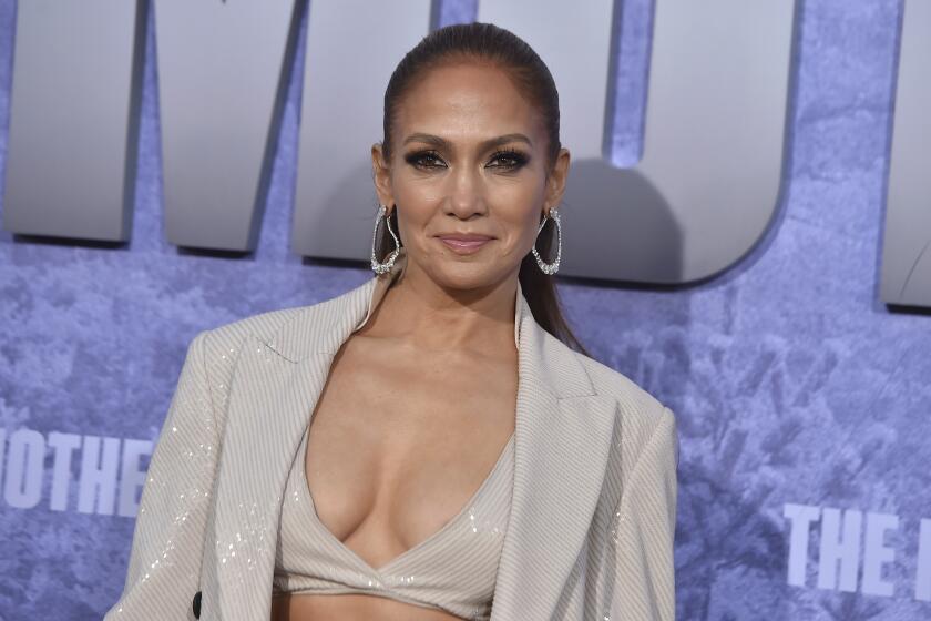 Jennifer Lopez posing in a sparkly suit, bra and earrings against a purple background.