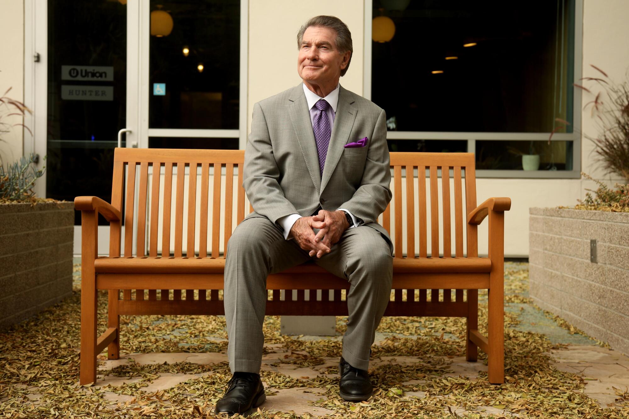 Former Dodgers first baseman Steve Garvey sits on a wooden bench dresses in a gray suit with a purple tie