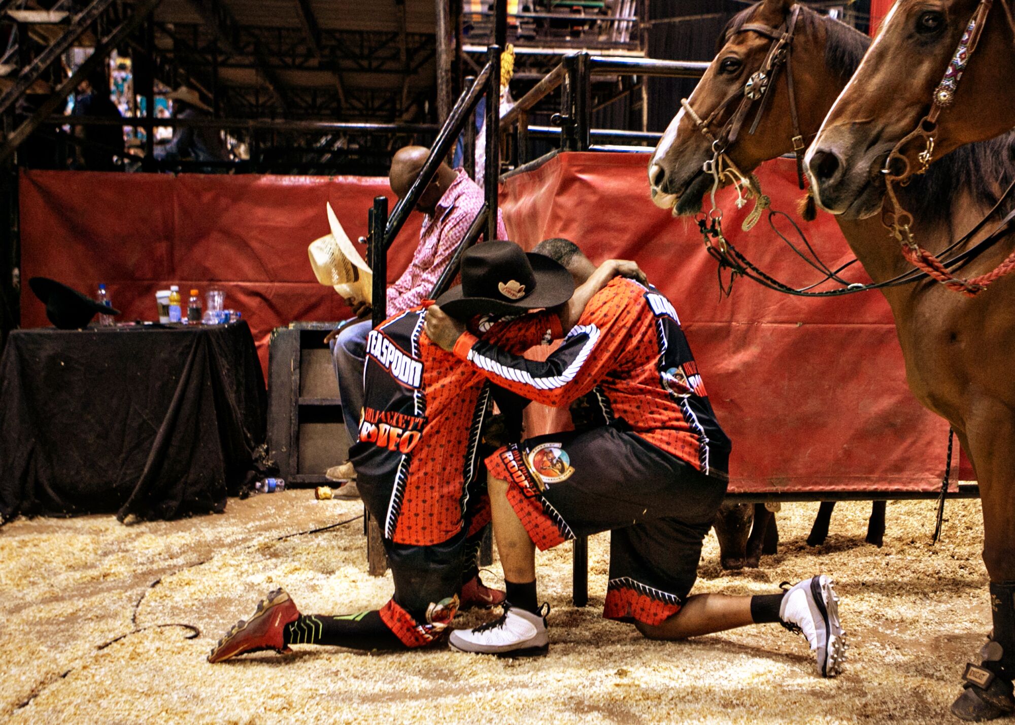Two men huddle and kneel in the sawdust beside two horses, as a man in the background removes his hat and bows his head.