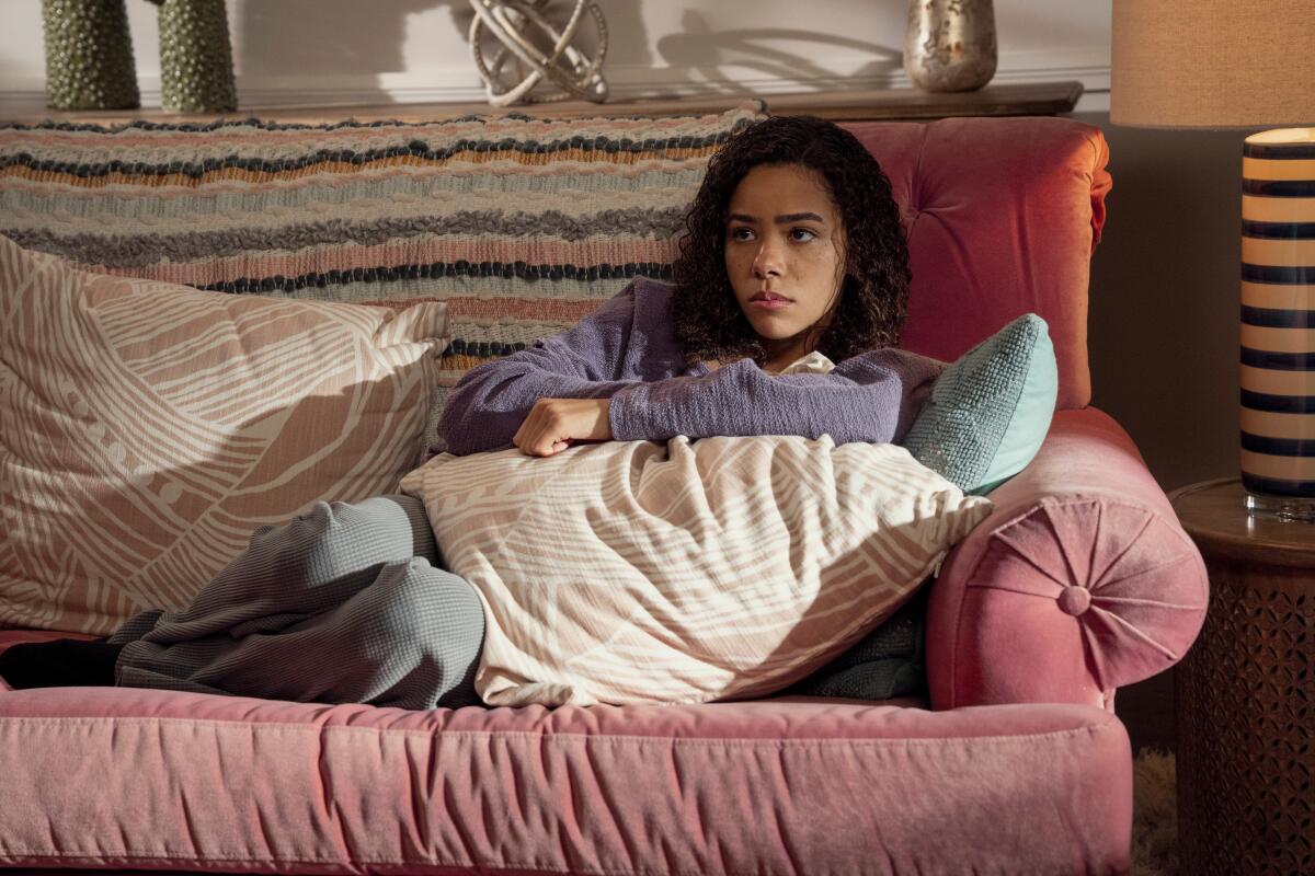 A teenage girl in a purple sweater sits on a pink couch with her legs curled to the side and her arms resting on a pillow.