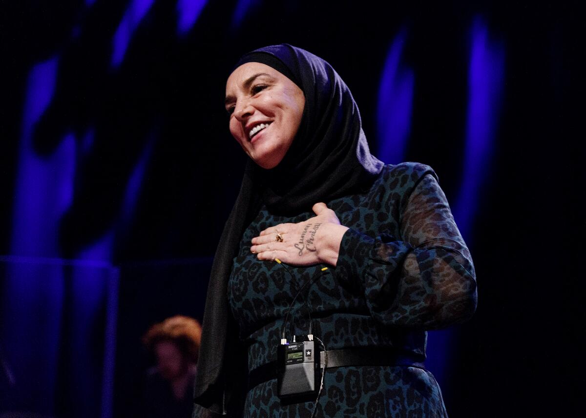 Singer-songwriter, wearing a head scarf, holds her hand to heart while standing on a stage. 