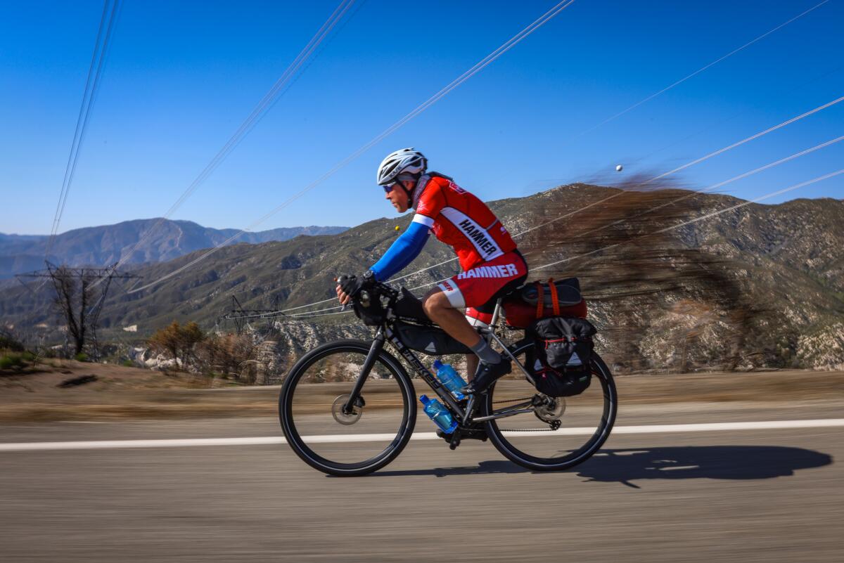 A man in a red bike racing outfit rides a bicycle on a road past hills.