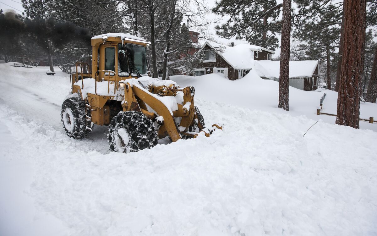 A snowplow clears a path on a snowy road. In the background are trees and a house with drifts piled to the windows.