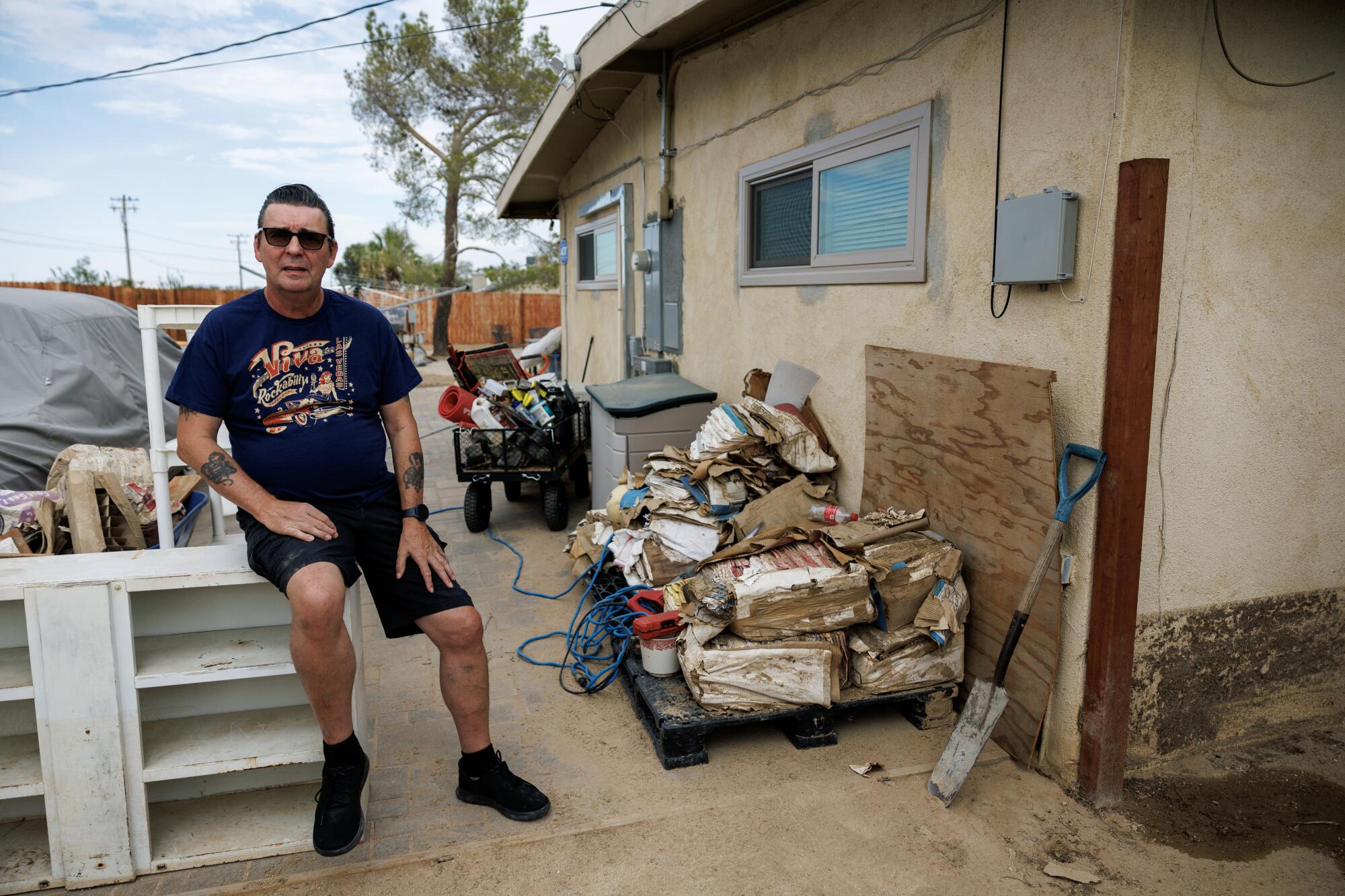 A man with dark hair, in sunglasses, dark shirt and shorts, sits outside a building with brown packages piled against a wall