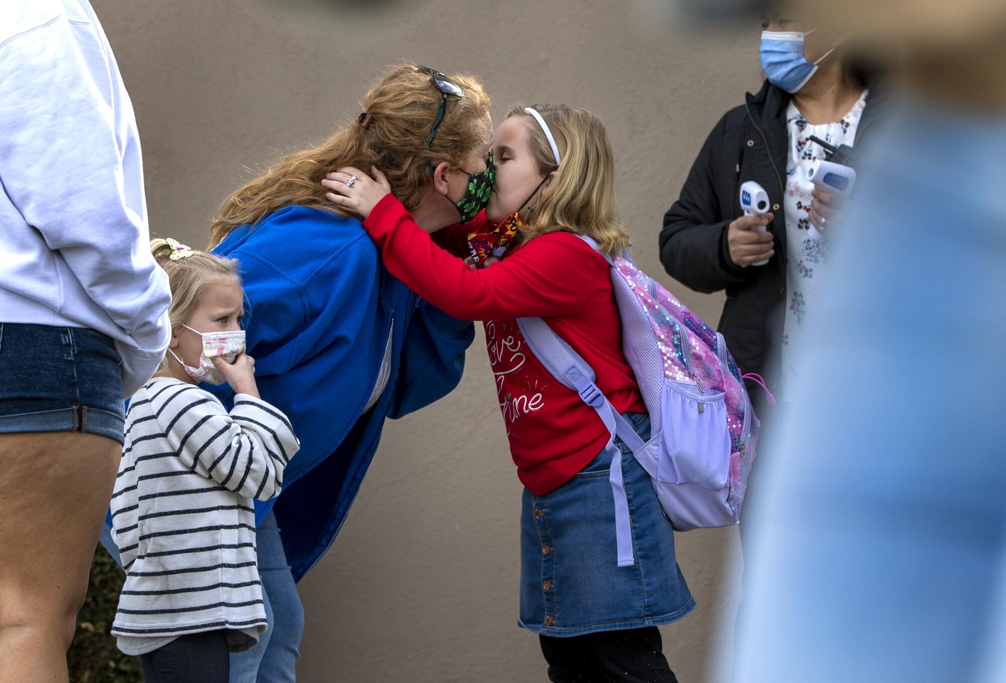 A young girl with a backpack kisses her mom on the mask. 