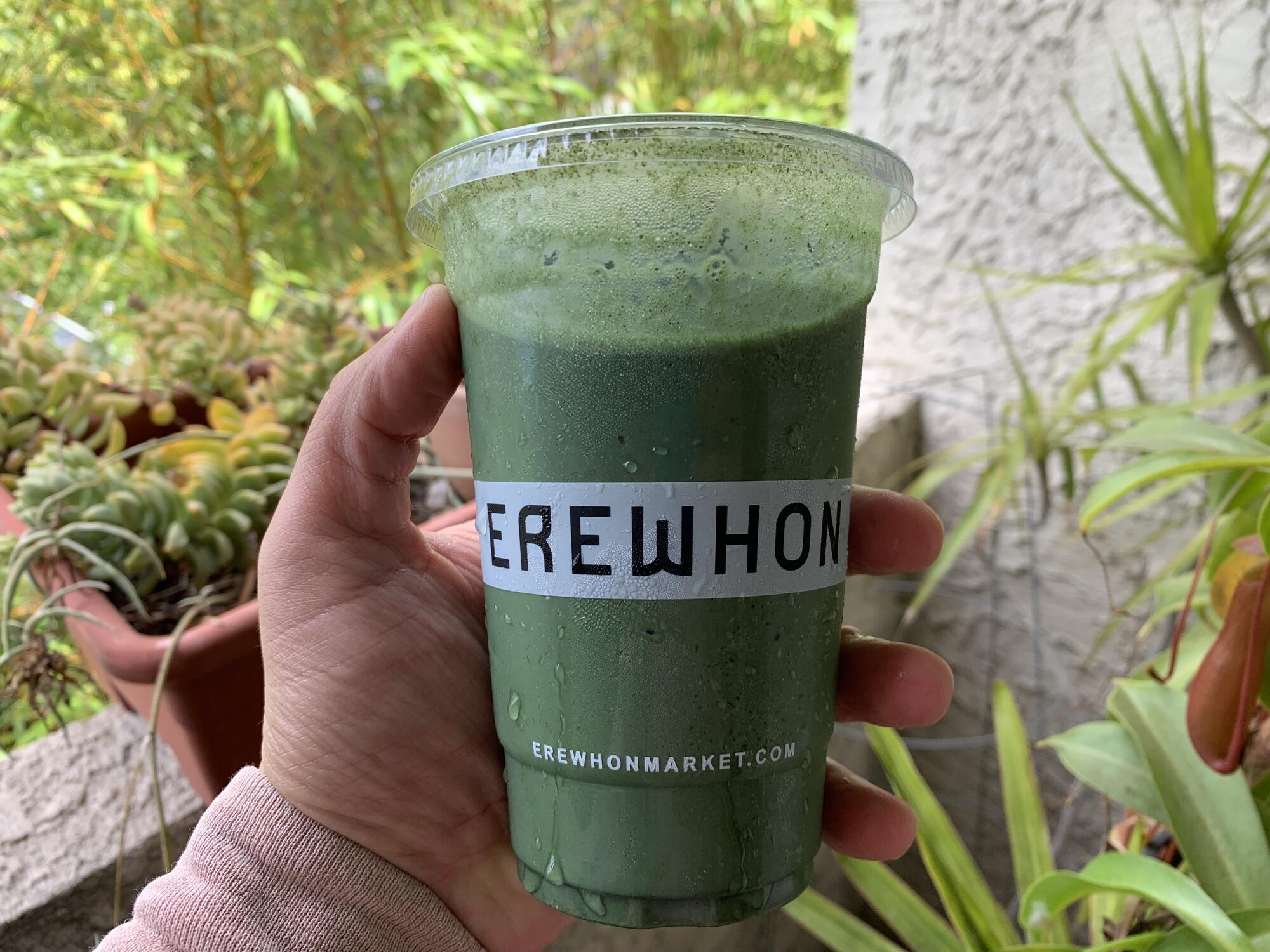 A hand holds a green smoothie in a plastic cup that says Erewhon.