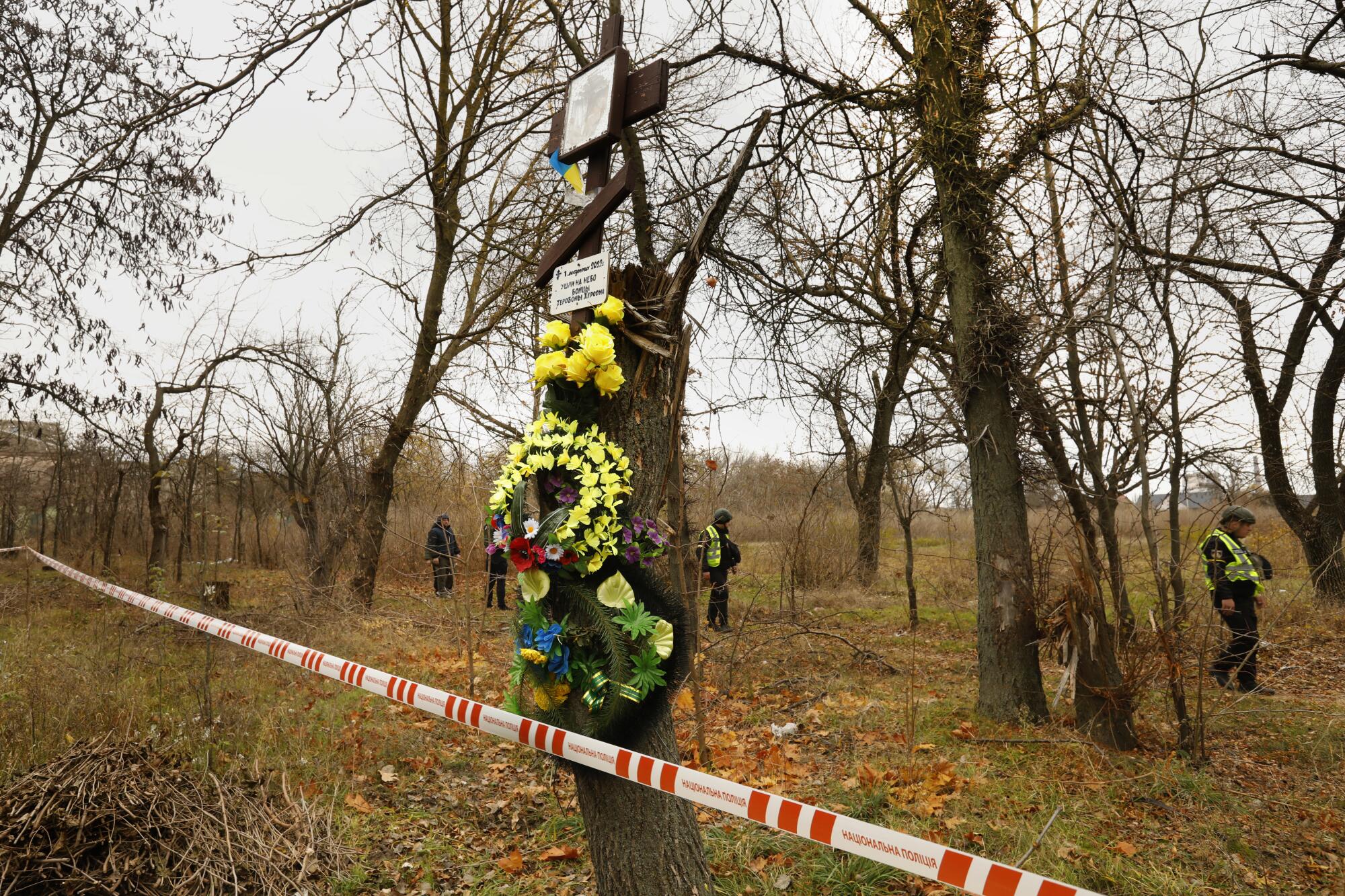 People in dark clothing walk among bare trees, one adorned with flowers and wreaths, cordoned off with red-and-white tape 