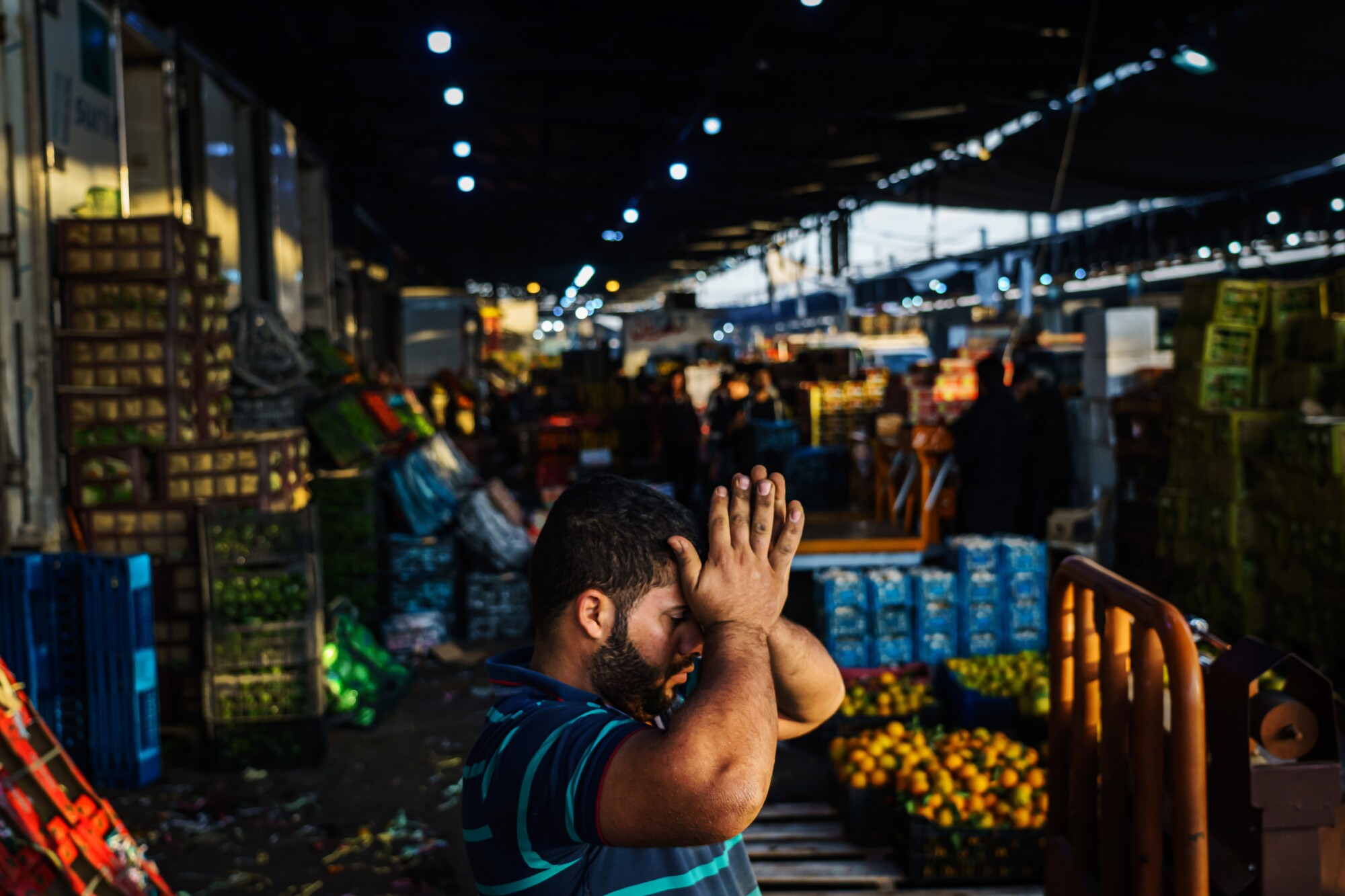 A side view of a man in a striped shirt holding his palms together near his forehead, surrounded by crates of produce
