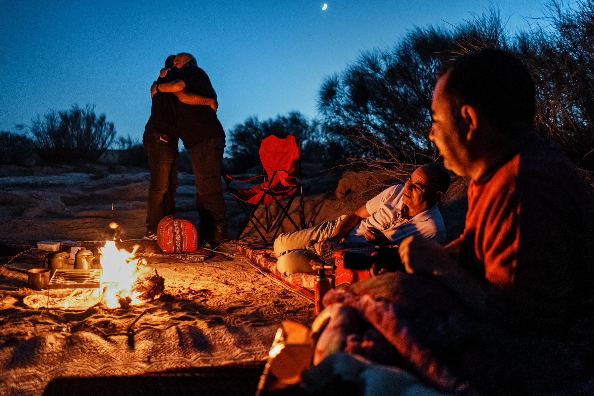 Two people embrace each near a campfire in the desert as others look on.