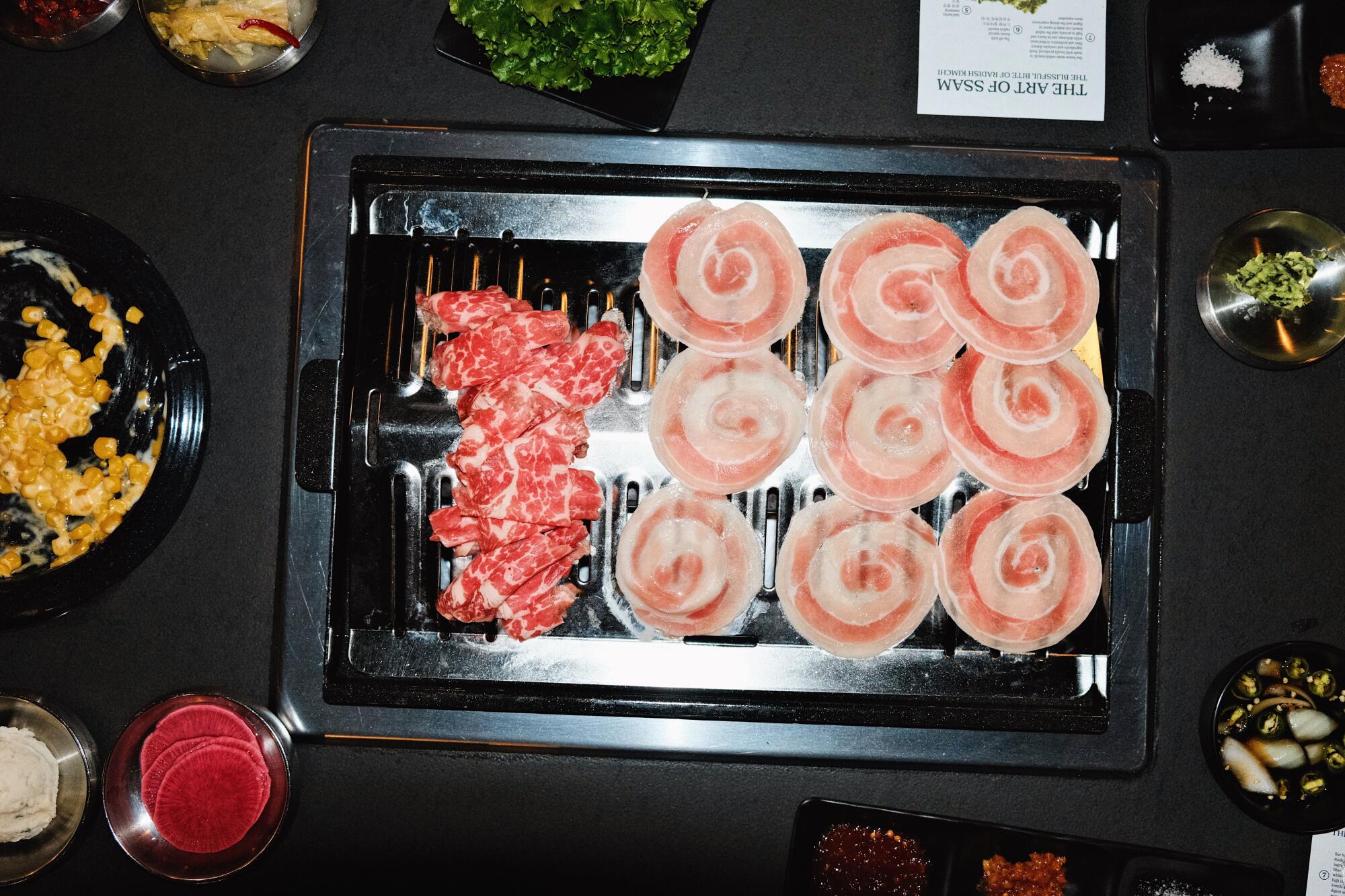 An overhead photo of a grill at Origin Korean BBQ: half is brisket, the other is pork belly disks. Banchan surround it.
