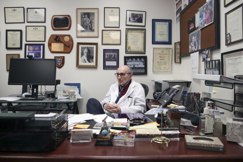 Dr. George Berci sits at a desk, before a wall of degrees and mementos