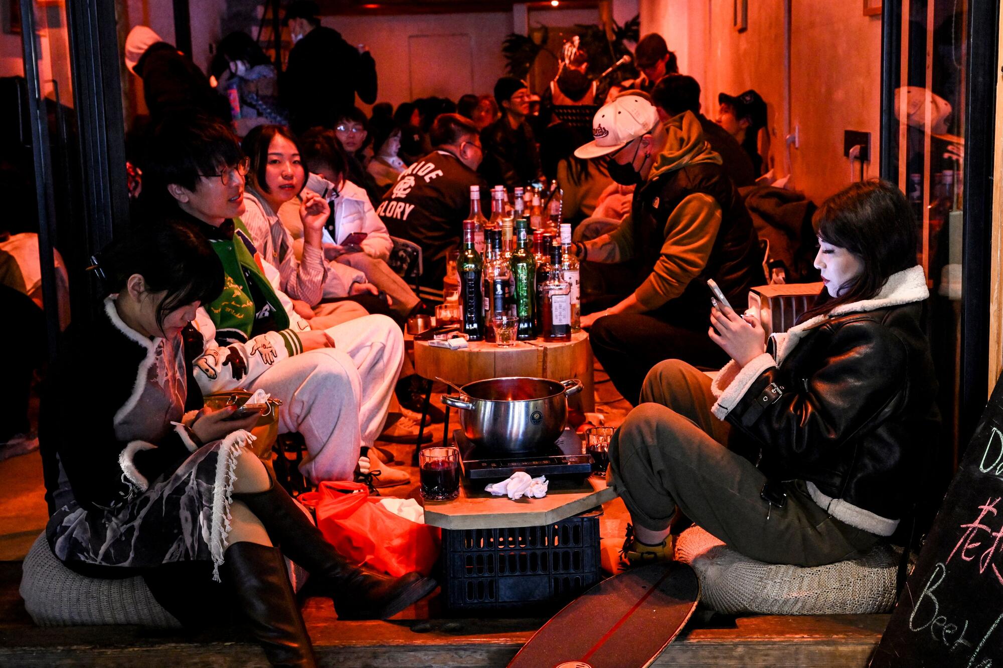 People sitting on cushions around low tables with bottles or a pot at a bar.